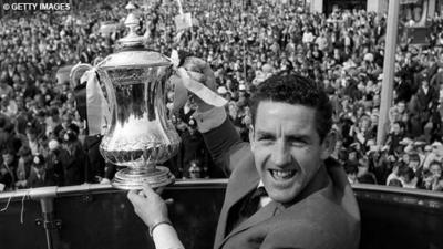Black and white image of Dave MacKay, facing the camera, holding aloft the FA Cup over a handrail. Below, a crowd of fans.