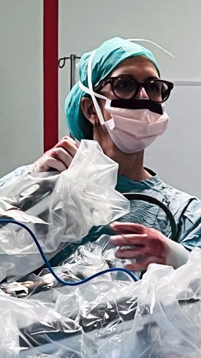 A surgeon in a mask with protective clothing on is holding onto the arms of a surgical robot. She is wearing gloves which are stained red with blood. She has glasses on and is looking away from the robot to the right of the screen.