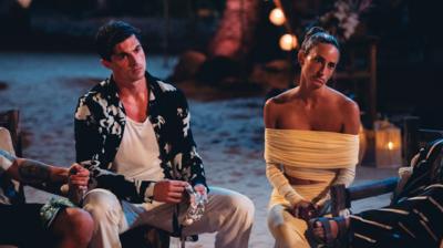 Oliver and Megan sitting around a fire with other cast members, looking frustrated