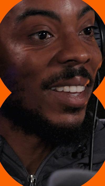Big Manny smiles in a radio studio wearing headphones. He has black facial hair and wears a black jacket