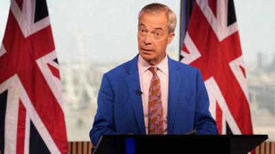 Nigel Farage stands behind a podium with two Union Jack's on each side
