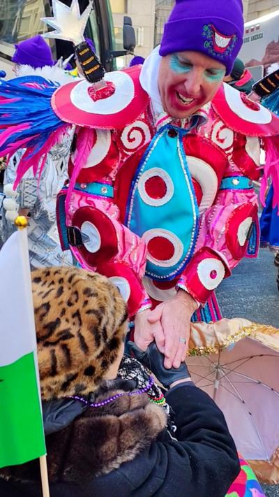 A woman smiles at the camera. She is sat on a brown armchair and hold a newspaper up to the camera. She is also holding a gold umbrella.
