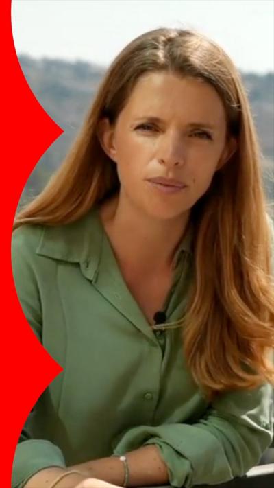 A woman with long dark blonde hair wearing a green blouselooks at the camera