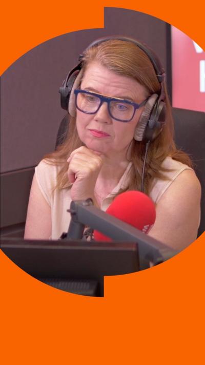 A woman with shoulder-length light ginger hair sits in front of a microphone and looks at someone off-screen