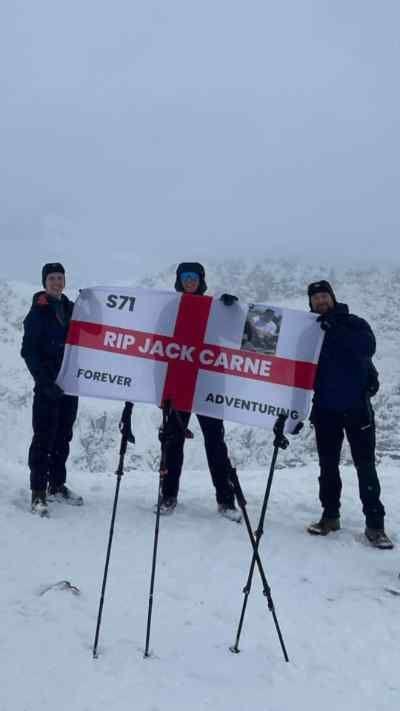 Three men on a snowy mountain hold up an England flag with a tribute on it