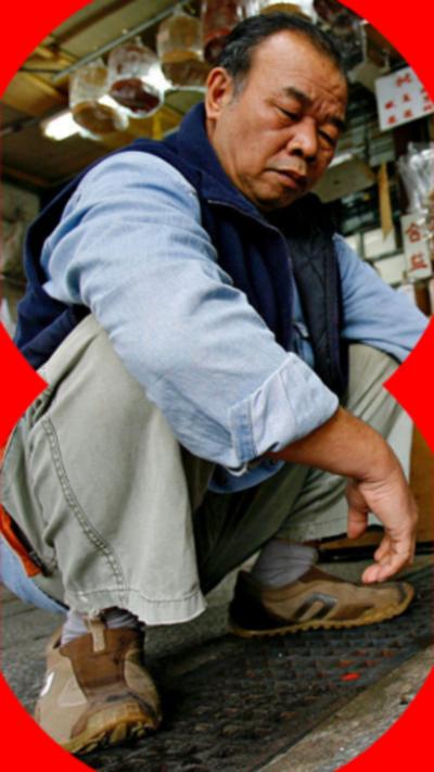 A man sits in the position of an 'Asian squat'. He's wearing a blue shirt and green trousers.