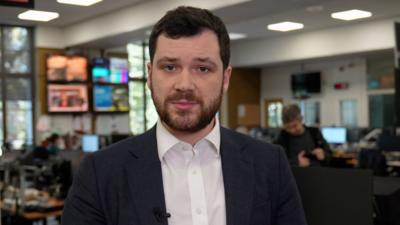 Henry Zeffman in BBC newsroom. He wears a shirt and blazer and has a beard.