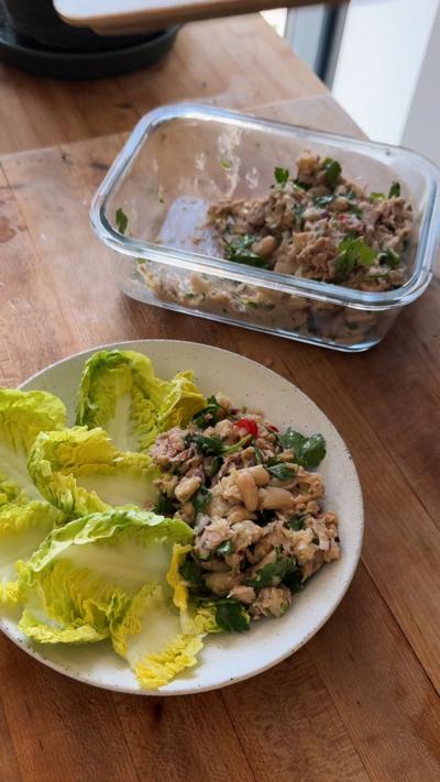 A glass lunchbox of a butter bean and tuna mix, white a portion on a plate with lettuce on a wooden surface. 