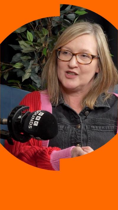 A woman with shoulder-length blonde hair sits in front of a microphone and talks to someone off camera.