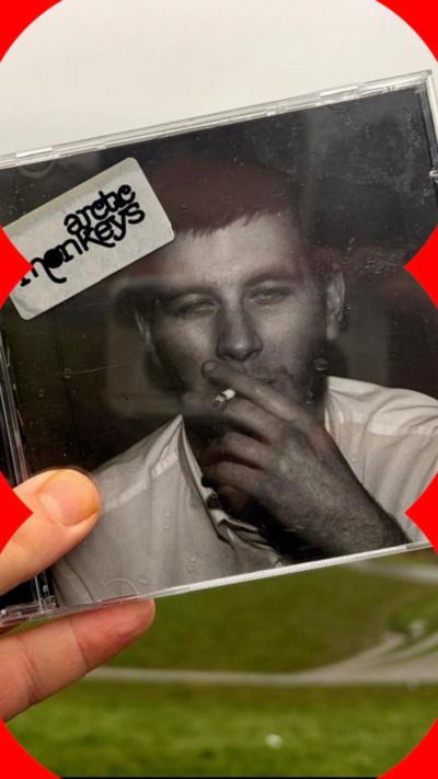 A hand holding up a CD of Arctic Monkeys' first album, featuring a black and white photo of a man smoking.