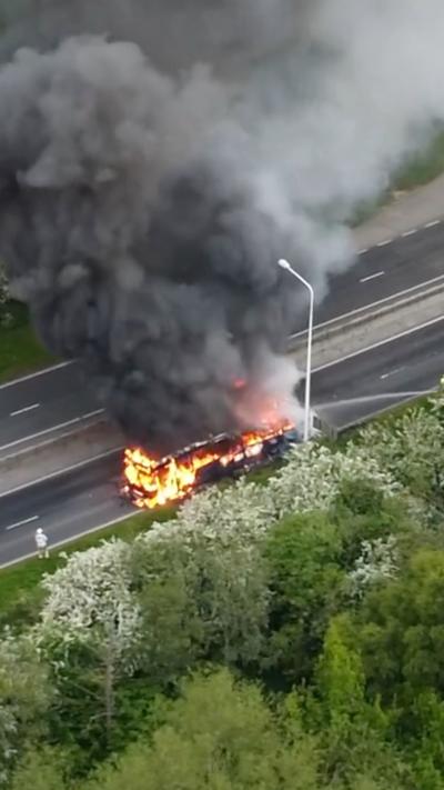 An aerial shot of a bus on fire. There are plumes of black smoke billowing into the air. Flames have engulfed the bus and firefighters are standing around it with a hose spraying water on to it.