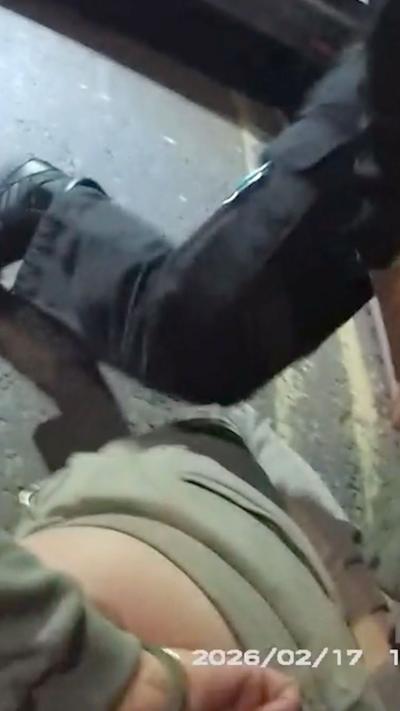 A man is handcuffed with his arms behind his back while a leg in black trousers is stood over him.