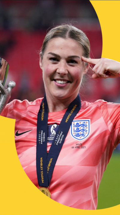 Mary Earps in a pink England shirt holding a trophy and wearing a medal pointing at her head with her hand
