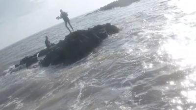 The silhouette of two children on a rock in the middle of a choppy sea. One is sitting down while the other stands. They are clearly stranded.