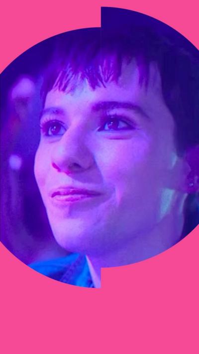 Byron stands in a club smiling. They have short brown hair and wear a blue denim shirt