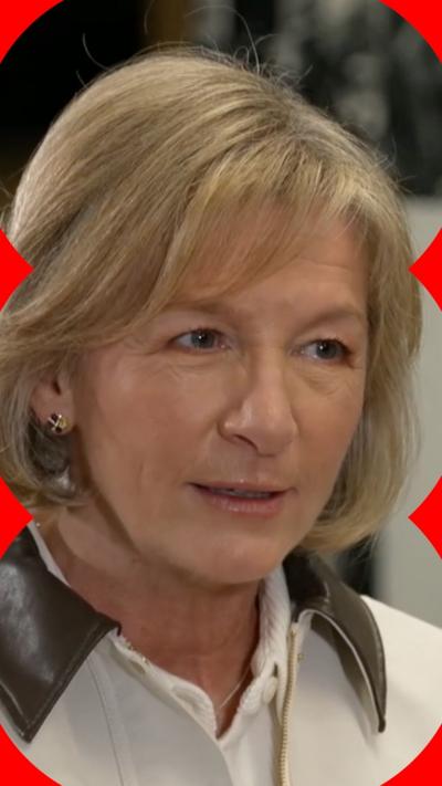 A woman with shoulder length blonde hair, wearing a white jacket with a brown collar