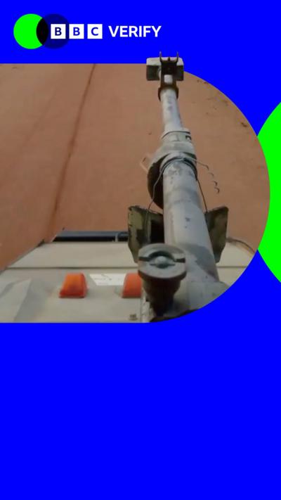 A tank cannon is seen as the vehicle drives along a dirt highway in Mali. The image is imposed over the BBC Verify colours and branding. 