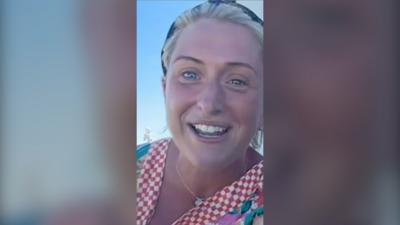 Laura Kenny smiles while speaking outside. She has blonde hair tied back with a headband, and wears a red and white checked top