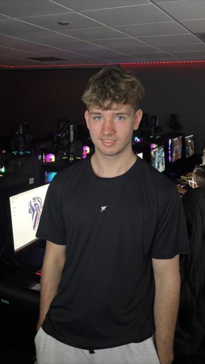 A teenage boy stands in the centre of the frame staring directly at the camera. He is wearing a black shirt and has blonde hair. Behind him are rows of gaming computers, glowing with a range of coloured lights. There are students in chairs playing games on these computers. 