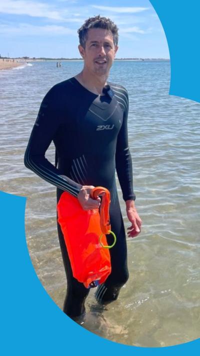 A man with short dark hair is wearing a full-body wetsuit and standing in the sea on a sunny day. He is holding an orange tow float.