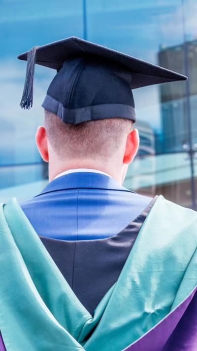 A stock photo from behind of a man with very short hair wearing a mortarboard hat and a teal coloured university gown over a blue suit