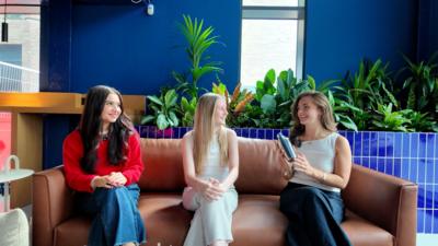 Three students sat on a brown sofa. The student on the right is showing the other two a cocktail shaker