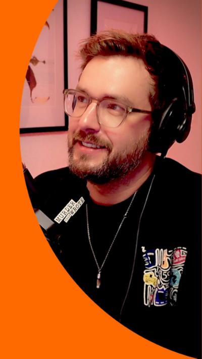 Iain Stirling wears black headphones and sits in-front of a mic and smiles. He wears a black long-sleeve jumper.