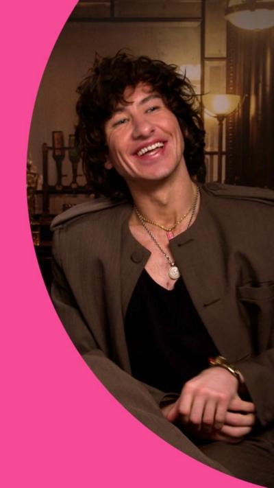 A man, Barry Keoghan, smiles. He has long wavy hair and wears a brown jacket.