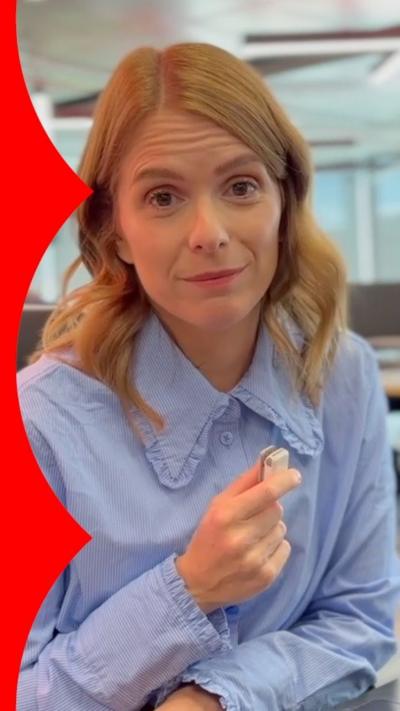 A woman with wavy ginger hair is sitting down. She is a wearing a blue blouse and holding a small microphone in front of her.
