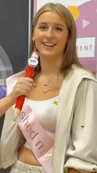 A girl wearing a pink sash and beige jumper smiles at the camera.