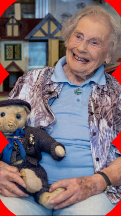 An elderly woman smiles while holding a vintage teddy bear in front of a doll's house display. 