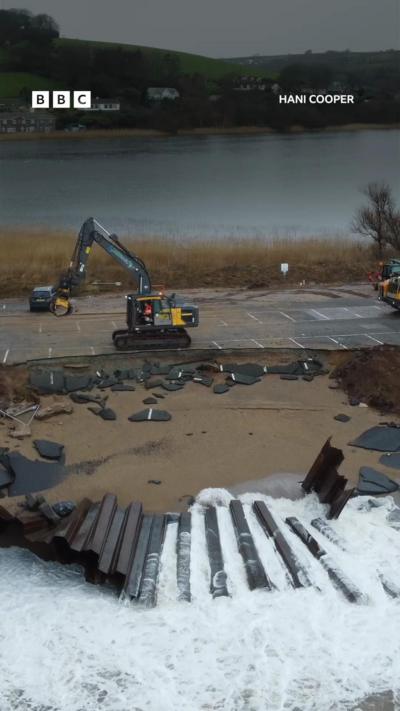 A digger is seen to the left of frame on land where a road once stood. The remnants of the road can be seen in the foreground on the sand with the sea in background.