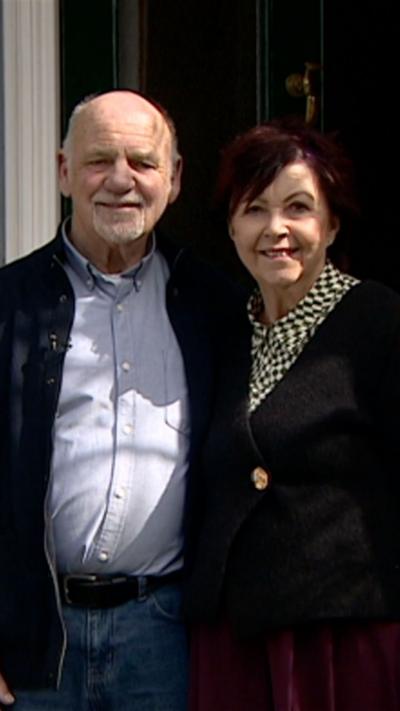 A man and woman stood together outside their black front door. The man is bald and has a grey goatie beard. He is wearing jeans, a blue jacket and blue shirt. The woman has short, brown hair and is wearing a dark top with a blue and yellow patterned shirt