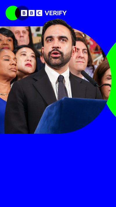Zorhan Mamdani wears a suit and is speaking into a microphone in front of a crowd of people 