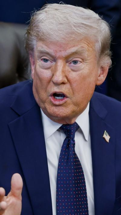 President Donald Trump wears a blue suit and blue tie as he speaks from a brown leather chair in the Oval Office.