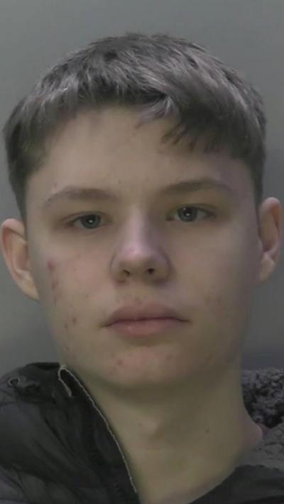 A mugshot of a boy with short brown hair and a black coat.