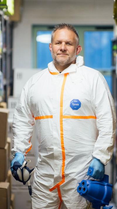 Up close photograph of Ben Giles. His is aged 49, has grey short hair. He wears a face mask around his neck. You can see his PPE around his neck, which is a hooded white suit.