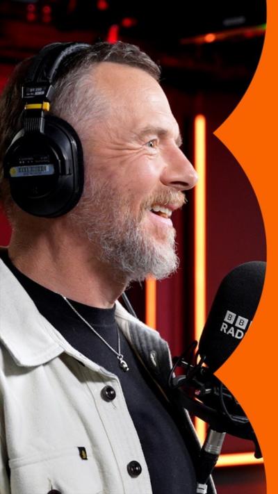 A man with short hair wearing headphones smiles at someone off-camera as he talks into a microphone