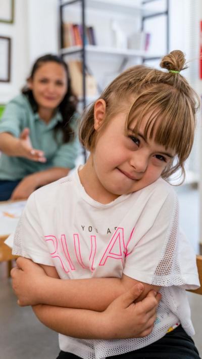 Young girl is close to camera with her arms crossed wearing a white t-shirt, an adult sits behind her at a low table in a modern looking room