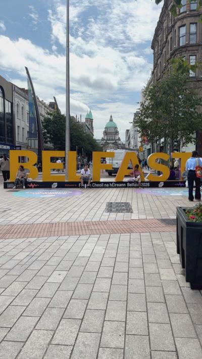 A big yellow lettered sign reading ' Belfast' in the city centre, placed with City Hall in the background.