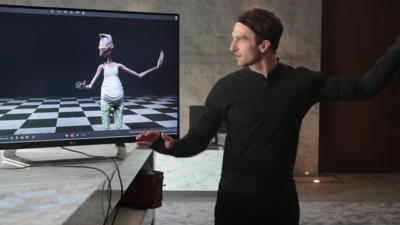 A man looks at a computer character on a screen while wearing a motion capture suit