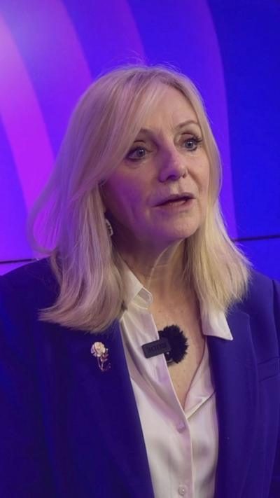 A woman wearing a purple blazer with mid length blonde hair stands in front of several tv screens