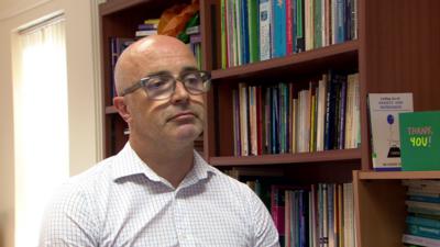 A bald man sits in front of brown book shelves filled with books. He wears a white and blue chequered shirt 