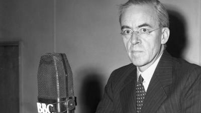 Black and white image of Sir Stafford Cripps in front of a microphone with a 91ȱ logo on it., looking at the camera.  He is wearing spectacles, a black pinstripe jack, a white shirt and a dark tie with white spots on it.