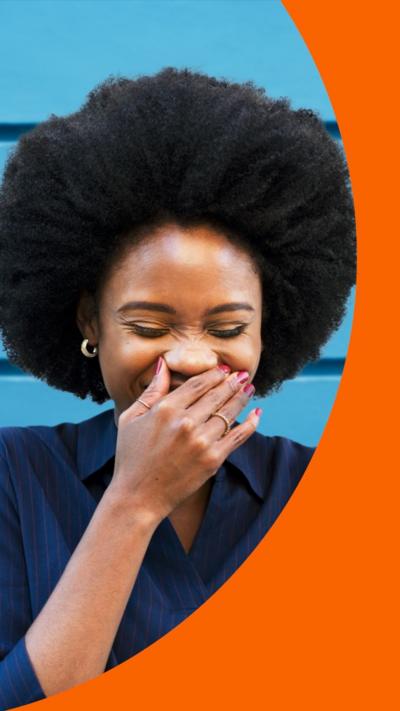 A woman covers her mouth as she laughs with her eyes closed, while standing in front of a blue wall