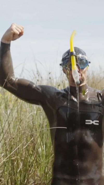 A man, dripping with water, in a grey wetsuit with a yellow snorkel and goggles raises his arms while waist deep in a brown bog.