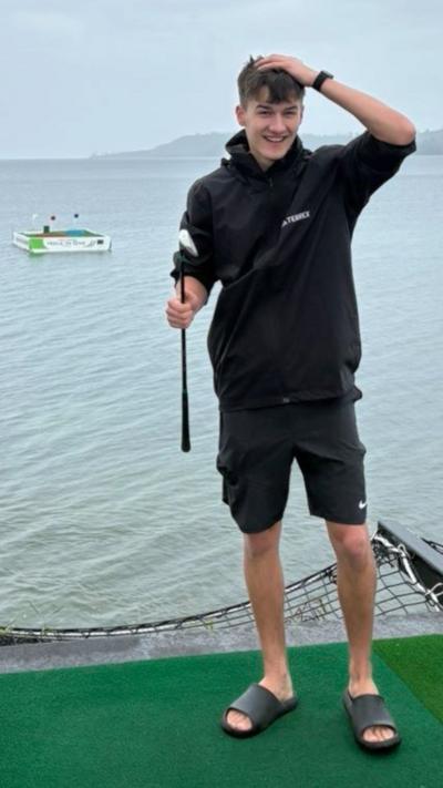 Teenage boy with golf club in one hand. His other hand is on his head in surprise. A floating golf hole is in the background.