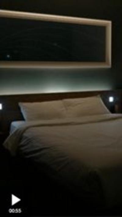 A bed in a darkened hotel room. The bed has white covers and sheets, and there is a white light coming from behind the headboard.