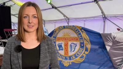 Carolyn Groat, neach-gairm Mòd Loch Abair, na seasamh air beulach bràtach a' Mhòid. 