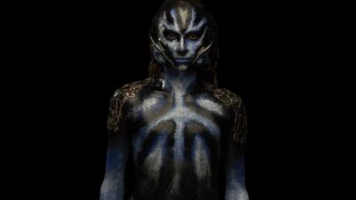 Model with black and blue make-up on their face and body, with prosthetic ears attached to their face.